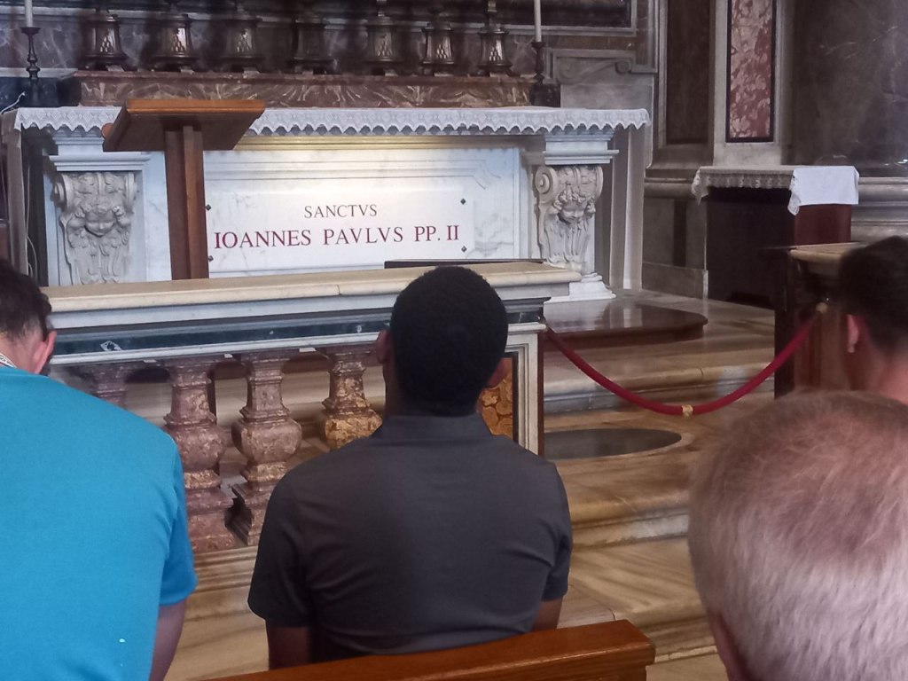 Personas sentadas en un banco de la iglesia frente al altar con el nombre 'IOANNES PAULUS PP. II' visible en la parte frontal.